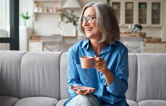 Happy relaxed mature older adult grey-haired woman drinking coffee relaxing on sofa at home